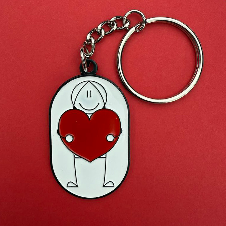 Foto van een sleutelhanger. Een metalen sleutelhanger met een getekend figuurtje dat een grote hart vasthoudt. Denk aan jou.